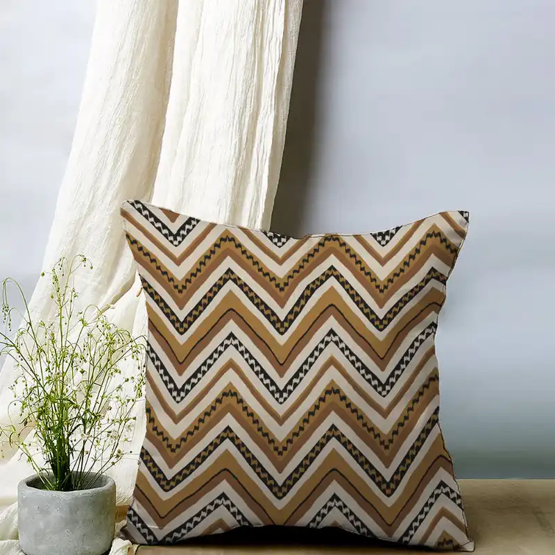 Decorative cushion with geometric chevron pattern in brown, black, and white, placed next to a white curtain and a plant in a grey pot.