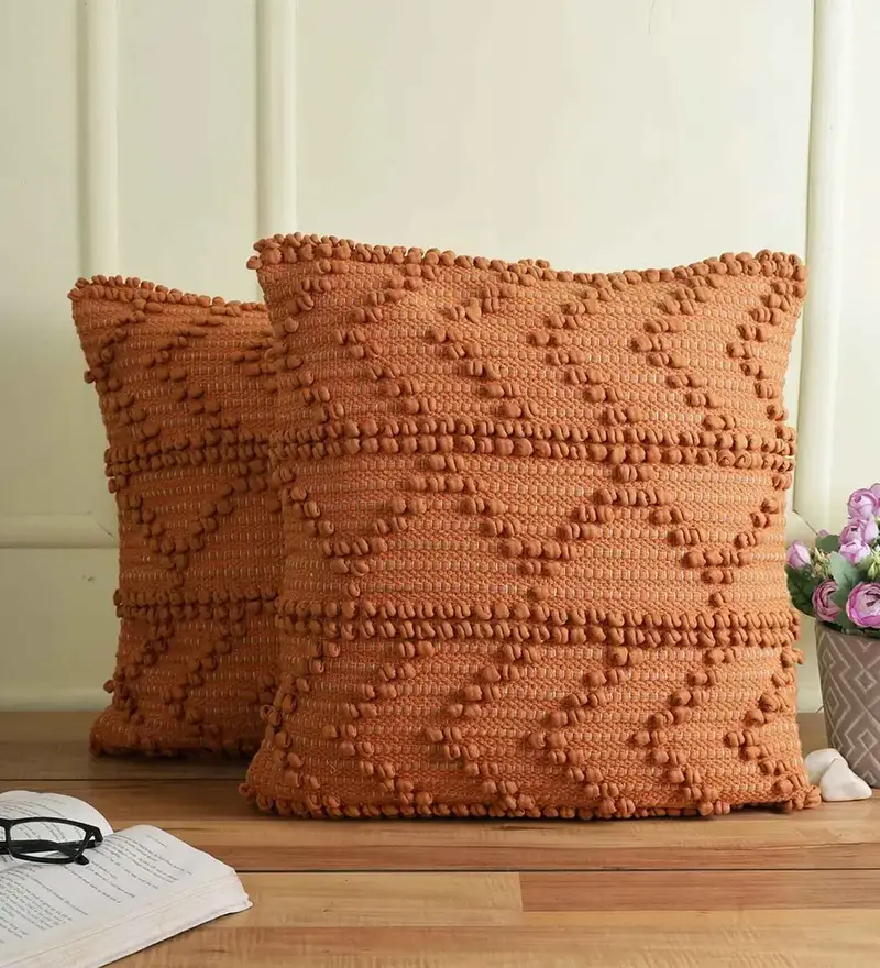 Two orange textured cushion pads with a zigzag pattern and pom-pom design, placed on a wooden surface next to a bouquet of flowers and an open notebook with reading glasses.