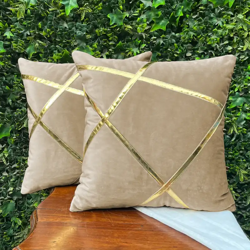 Two beige velvet cushion pads with gold geometric patterns placed on a wooden surface against a green leafy backdrop.