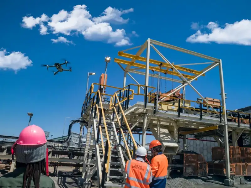 Construction workers observe a drone flying near a construction site, highlighting the use of drones in construction projects under a clear blue sky.