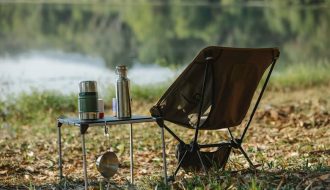 A peaceful lakeside campsite with a folding chair, table, thermoses, and saucepan.