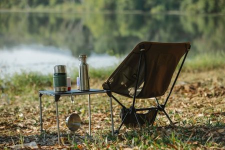 A peaceful lakeside campsite with a folding chair, table, thermoses, and saucepan.