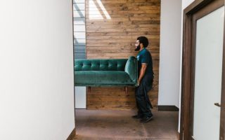 A mover carrying a green sofa through a hallway with a wooden wall background.