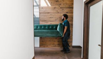 A mover carrying a green sofa through a hallway with a wooden wall background.