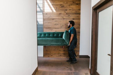 A mover carrying a green sofa through a hallway with a wooden wall background.
