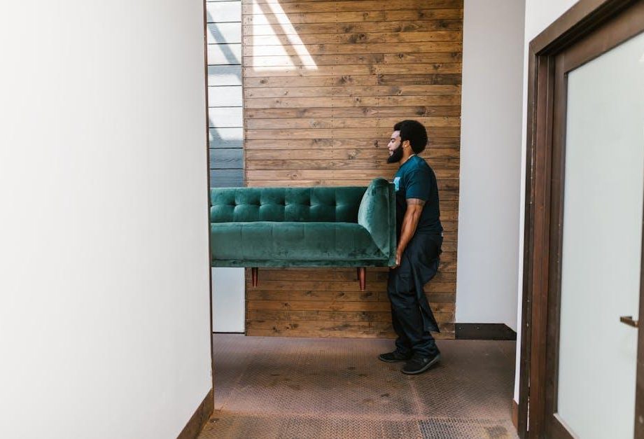 A mover carrying a green sofa through a hallway with a wooden wall background.