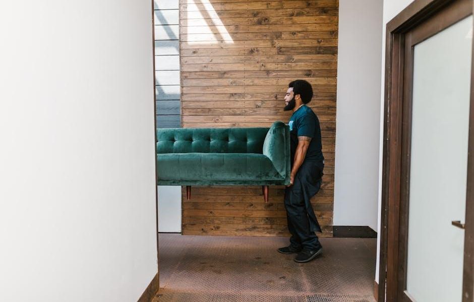 A mover carrying a green sofa through a hallway with a wooden wall background.